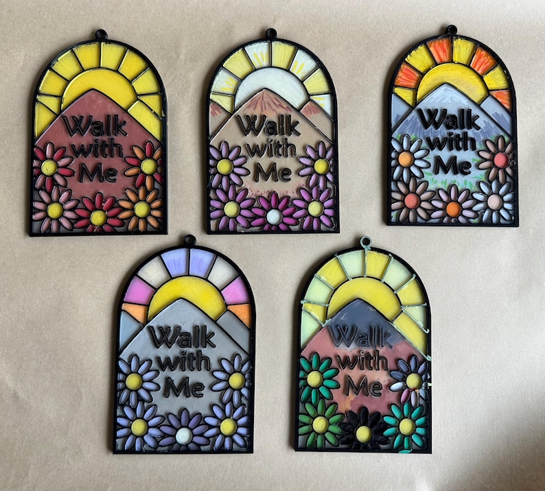 Puede incluir: Cinco colgantes de pared arqueados, estilo vidriera. Cada uno presenta una escena de monta&ntilde;a con un sol, flores y el texto "Walk with Me". Las paletas de colores incluyen tonos de amarillo, naranja, rosa, morado y verde, con bordes negros.