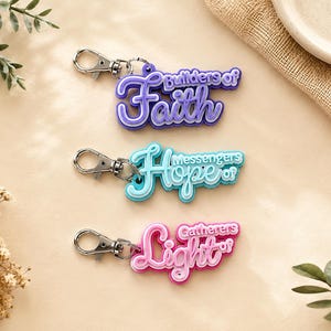 May include: Three layered acrylic keychains with silver-tone lobster clasps. The top keychain is purple with the text Builders of Faith. The middle keychain is teal with the text Messengers of Hope. The bottom keychain is pink with the text Gatherers of Light.
