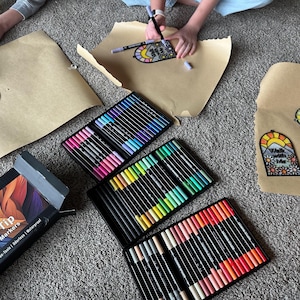Puede incluir: Vista a&eacute;rea de materiales de manualidades, que incluyen 72 rotuladores acr&iacute;licos de doble punta en varios colores y papel marr&oacute;n con dibujos. Los dibujos presentan un sol, monta&ntilde;as y el texto "Walk with Me".