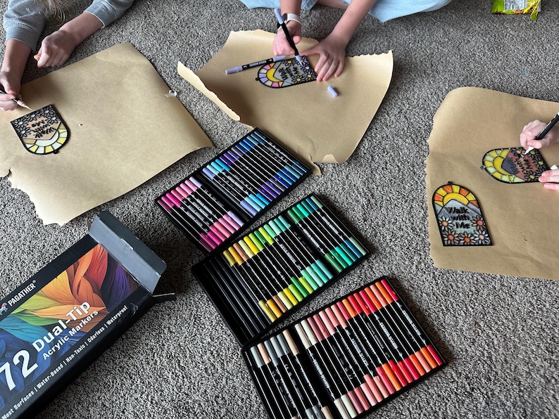 Puede incluir: Vista a&eacute;rea de materiales de manualidades, que incluyen 72 rotuladores acr&iacute;licos de doble punta en varios colores y papel marr&oacute;n con dibujos. Los dibujos presentan un sol, monta&ntilde;as y el texto "Walk with Me".