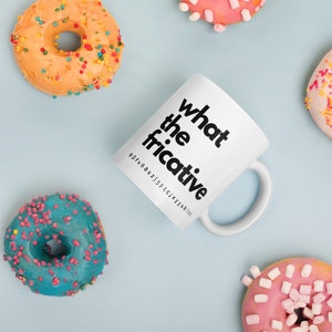Puede incluir: Taza de cerámica blanca con texto negro que dice "what the fricative". La taza está rodeada de donuts coloridos con chispas.