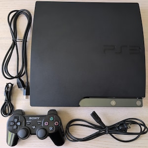 May include: A black Sony PlayStation 3 game console featuring the PS3 logo embossed on the top surface. The set includes a black wireless controller with Sony branding, a black power cable, and a black HDMI cable, all presented as a complete gaming unit.