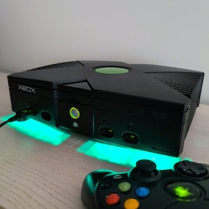 May include: A black original Xbox video game console featuring a prominent X-shaped top design with a green circular emblem. The front panel displays the Xbox logo, a power button with a glowing green ring, and controller ports. Bright green light emanates from beneath the console, accompanied by a black game controller.