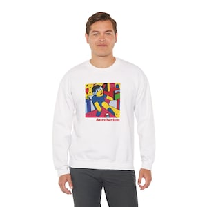 May include: White sweatshirt featuring a colorful graphic of a figure in a cubist style. The graphic includes blue, yellow, red, and black elements, with the word "Aucubetism" in red below the image. The sweatshirt is a casual, long-sleeved garment.