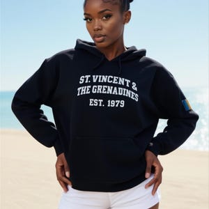 May include: Black hooded sweatshirt with the text "ST. VINCENT & THE GRENADINES EST. 1979" in white. The sweatshirt has a front pocket and a small flag detail on the sleeve. The model is wearing white shorts.