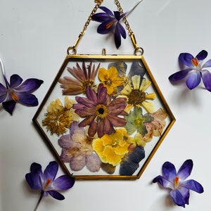 May include: A hexagonal gold frame containing a pressed flower arrangement. The flowers are in shades of purple, yellow, and brown. The frame is hanging from a gold chain.