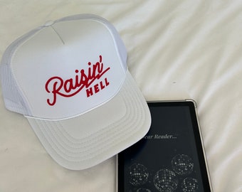 May include: White trucker hat with red embroidered text that reads "Raisin' Hell" on a white bed with a black Kindle e-reader.