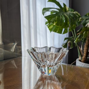 May include: A clear, faceted crystal bowl with a scalloped rim. The bowl has a square base that tapers upwards to the wider, open top. The bowl is sitting on a reflective surface, and the light refracts through the crystal.