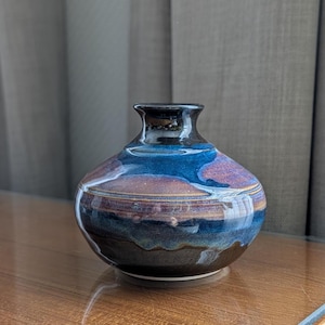 May include: A small ceramic vase with a rounded body and a narrow neck. The glaze features a blend of deep blue, dark brown, and muted purple tones, with horizontal bands of color that transition smoothly across the surface of the vessel.