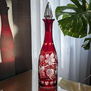 May include: A tall, ruby red glass decanter featuring a clear, pointed glass stopper. The body of the decanter is decorated with etched white designs of grape clusters and vine leaves, with a row of clear circular cutouts near the neck.