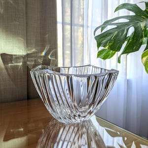 May include: A clear glass bowl featuring a distinct scalloped rim and deep vertical fluted ridges that create a textured, light-reflecting surface. The bowl has a wide, open top and a tapered base, showcasing a transparent, crystalline appearance.