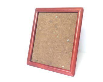 May include: A rectangular, reddish-brown wooden picture frame with a brown backing. The frame is angled slightly, showcasing its clean lines and simple design. The back has several small holes.