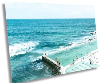 May include: A scenic photograph of a turquoise ocean meeting a light blue sky. A rectangular swimming pool is built into the ocean's edge, with people swimming and standing around the pool. The image is a print.