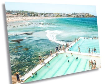 May include: A vibrant photograph of a beach scene with turquoise water, a sandy shore, and a swimming pool. People are swimming in the ocean and the pool. Buildings line the coast under a clear blue sky. The image is a print.