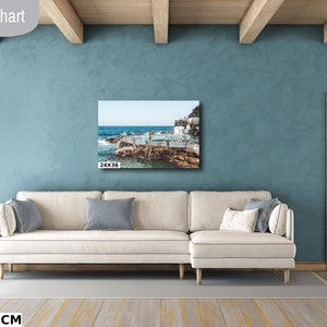 May include: A living room size chart showing a 24x36 inch canvas print of a seascape with a white building and people swimming in the ocean. The print is hanging on a light blue wall above a white couch.