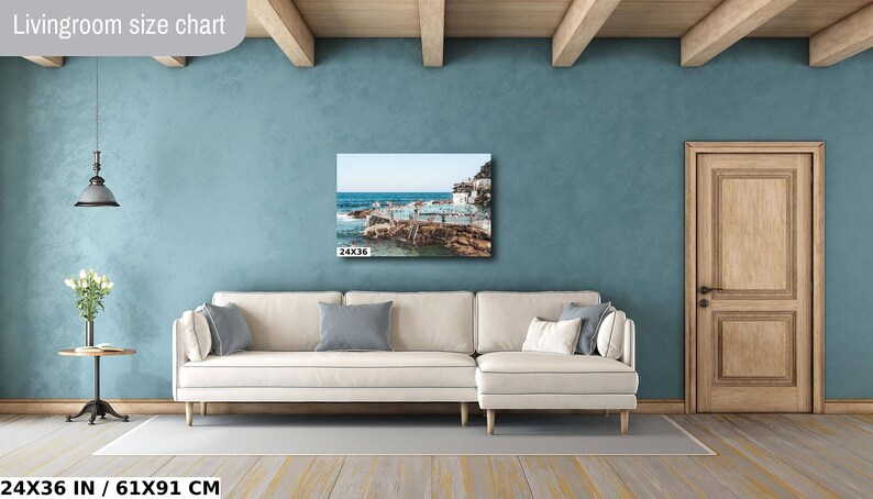 May include: A living room size chart showing a 24x36 inch canvas print of a seascape with a white building and people swimming in the ocean. The print is hanging on a light blue wall above a white couch.