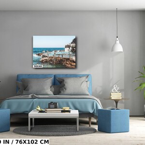May include: A blue bed with white pillows and a blue ottoman in front of it. A white lamp hangs from the ceiling. A framed photograph of a beach scene with people swimming in the water hangs on the wall above the bed. The photograph is 30x40 inches.