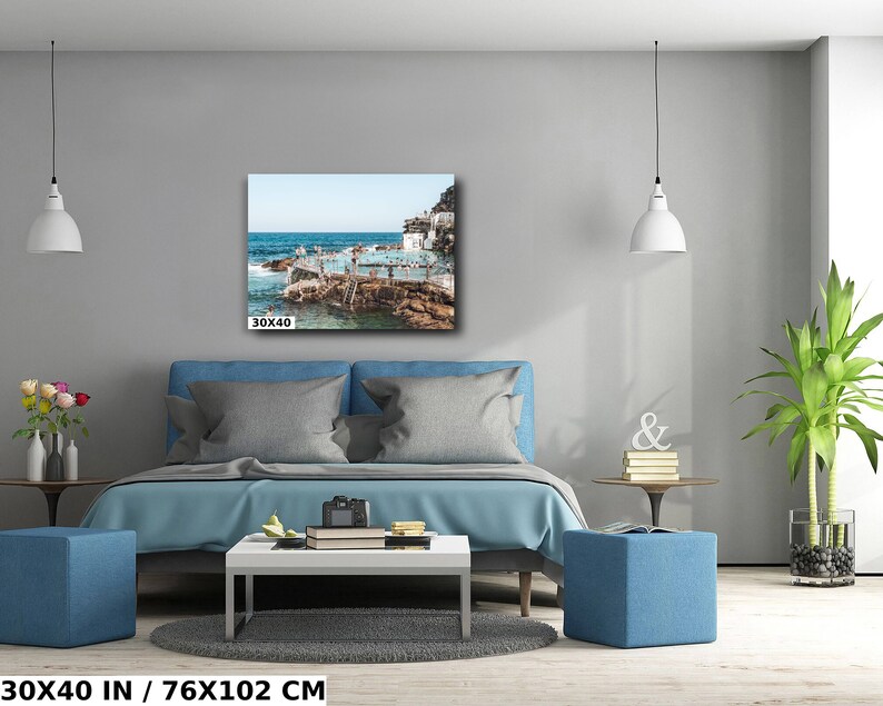May include: A blue bed with white pillows and a blue ottoman in front of it. A white lamp hangs from the ceiling. A framed photograph of a beach scene with people swimming in the water hangs on the wall above the bed. The photograph is 30x40 inches.