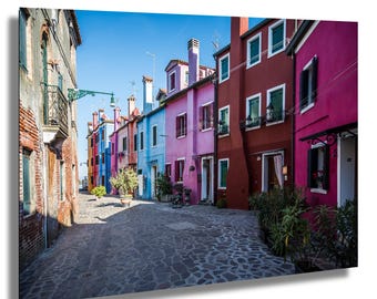 Può includere: Una stretta via acciottolata fiancheggiata da case colorate in varie tonalità di rosa, blu e rosso. Le case hanno più finestre e alcune hanno balconi. La strada è vuota tranne che per alcune piante.