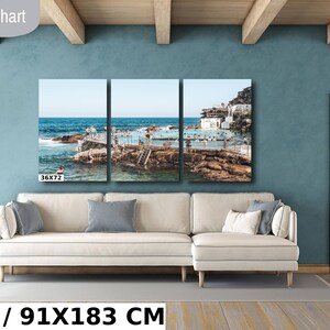 May include: A three-panel canvas print of a beach scene with a pool and people swimming. The print is 36x72 inches or 91x183 centimeters. The print is hanging on a wall in a living room with a white couch and a wooden door.
