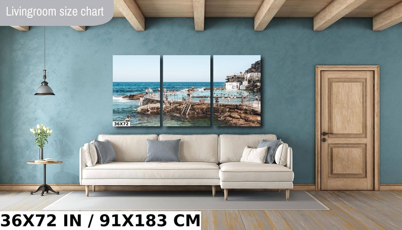May include: A three-panel canvas print of a beach scene with a pool and people swimming. The print is 36x72 inches or 91x183 centimeters. The print is hanging on a wall in a living room with a white couch and a wooden door.