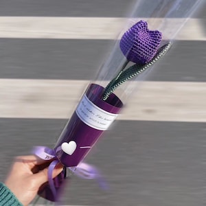 K&ouml;nnte beinhalten: Eine einzelne, handgefertigte lila H&auml;keltulpe in einem dekorativen lila Papierkegel. Der Kegel ist mit einer transparenten Plastikh&uuml;lle umwickelt und mit einem lila Band gebunden. Der Text auf dem Kegel lautet "Best wishes like heaven in general, which is sweet."