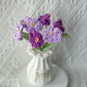 May include: A handmade crochet bouquet of purple and lavender flowers with yellow centres, displayed in a small white crocheted vase. The vase has a small label. The flowers have green stems and leaves. The background is a white textured fabric.