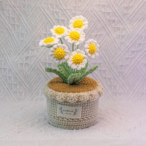 May include: A crocheted daisy flower arrangement in a beige pot. The daisies have white petals and yellow centres. The pot is beige with a scalloped edge and a small label that reads "Handmade with love."
