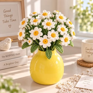 May include: A bouquet of crocheted daisy flowers with white petals and yellow centres, featuring green stems and leaves, arranged in a smooth, round, bright yellow ceramic vase. The flowers are crafted with detailed yarn stitching.