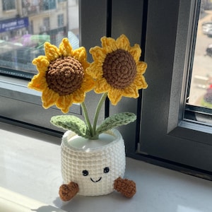 May include: A crocheted sunflower plant with two yellow flowers and brown centers. The plant is in a white pot with a smiling face and brown feet. The plant is placed near a window.
