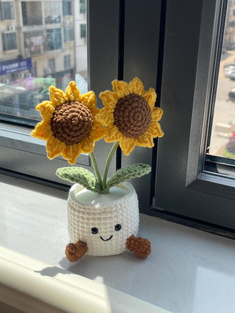 May include: A crocheted sunflower plant with two yellow flowers and brown centers. The plant is in a white pot with a smiling face and brown feet. The plant is placed near a window.