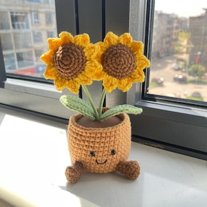 May include: A crocheted sunflower pot with two yellow sunflowers with brown centers. The pot is orange with a smiling face and small feet. The flowers have green leaves and are placed in front of a window.