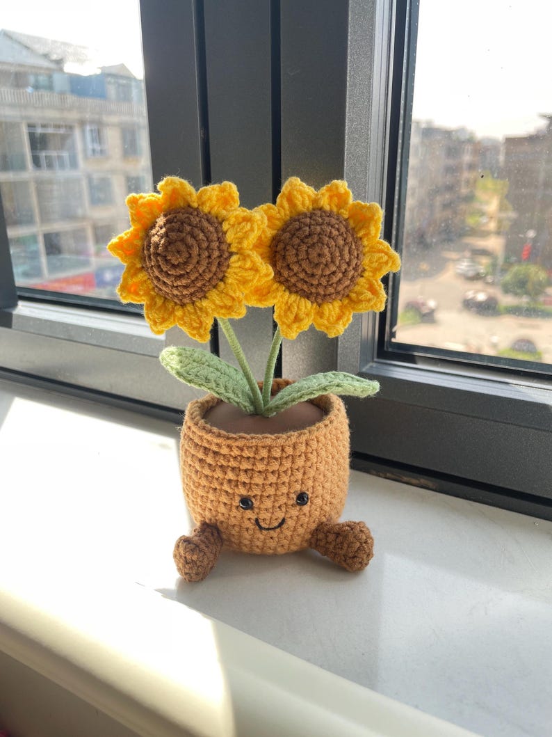 May include: A crocheted sunflower pot with two yellow sunflowers with brown centers. The pot is orange with a smiling face and small feet. The flowers have green leaves and are placed in front of a window.