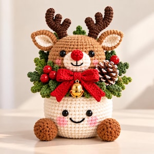 May include: A crocheted reindeer figurine with brown antlers, a red nose, and black eyes, nestled in a cream-colored crocheted pot with a smiling face. The reindeer is surrounded by green crocheted leaves, red berries, a pinecone, and a red bow with a gold bell.
