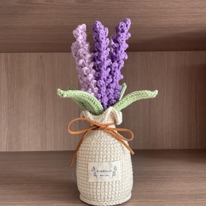 Può includere: Un bouquet di lavanda all'uncinetto in un vaso di colore crema all'uncinetto con un nastro marrone e un'etichetta che dice "Fatto a mano con amore".