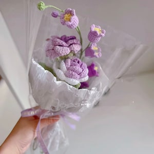 May include: A bouquet of crocheted flowers in shades of purple and white, wrapped in white tissue paper and tied with a purple ribbon.