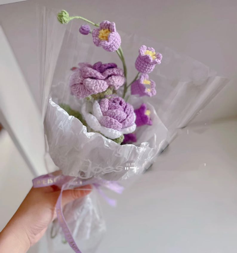 May include: A bouquet of crocheted flowers in shades of purple and white, wrapped in white tissue paper and tied with a purple ribbon.