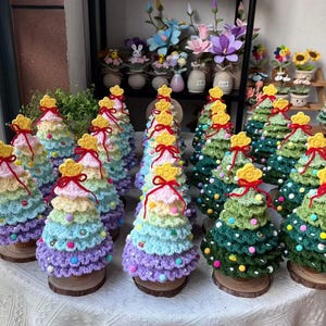 May include: A collection of small, cone-shaped crochet Christmas trees arranged on a table. Each tree features tiered, scalloped layers in pastel or green yarn, decorated with tiny colorful pom-poms, a red bow, and a yellow star topper on a wood base.