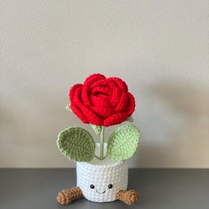 May include: A handmade crochet rose in a white pot with a smiling face and small brown feet. The red rose has green leaves and a green stem. The pot is approximately 4 inches tall.