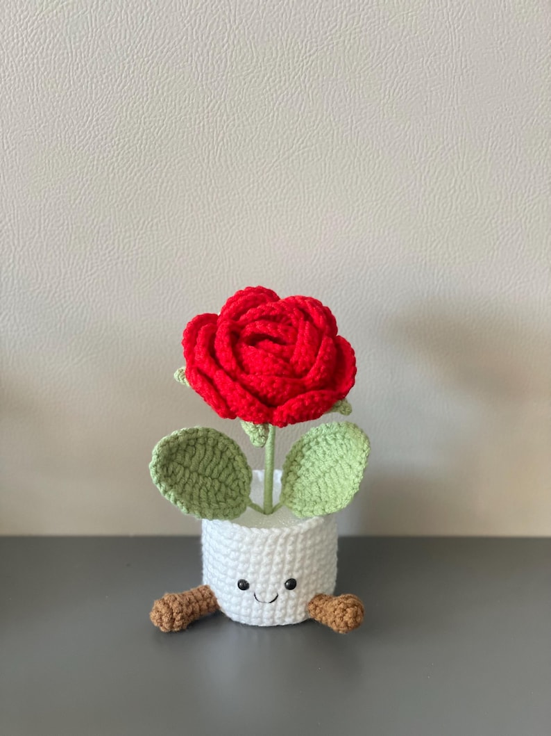 May include: A handmade crochet rose in a white pot with a smiling face and small brown feet. The red rose has green leaves and a green stem. The pot is approximately 4 inches tall.
