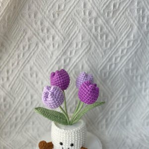 May include: A handmade crochet flower arrangement featuring four purple and lavender tulips in a white pot with a smiling face and brown feet. The arrangement includes green leaves and is set on a white surface.