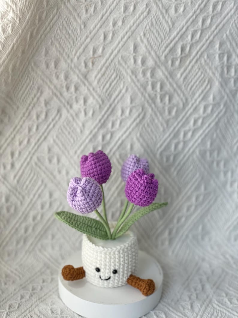 May include: A handmade crochet flower arrangement featuring four purple and lavender tulips in a white pot with a smiling face and brown feet. The arrangement includes green leaves and is set on a white surface.