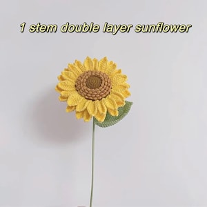 May include: A single-stem crocheted sunflower with a double layer of yellow petals and a brown center. The sunflower has a green leaf and a long green stem.