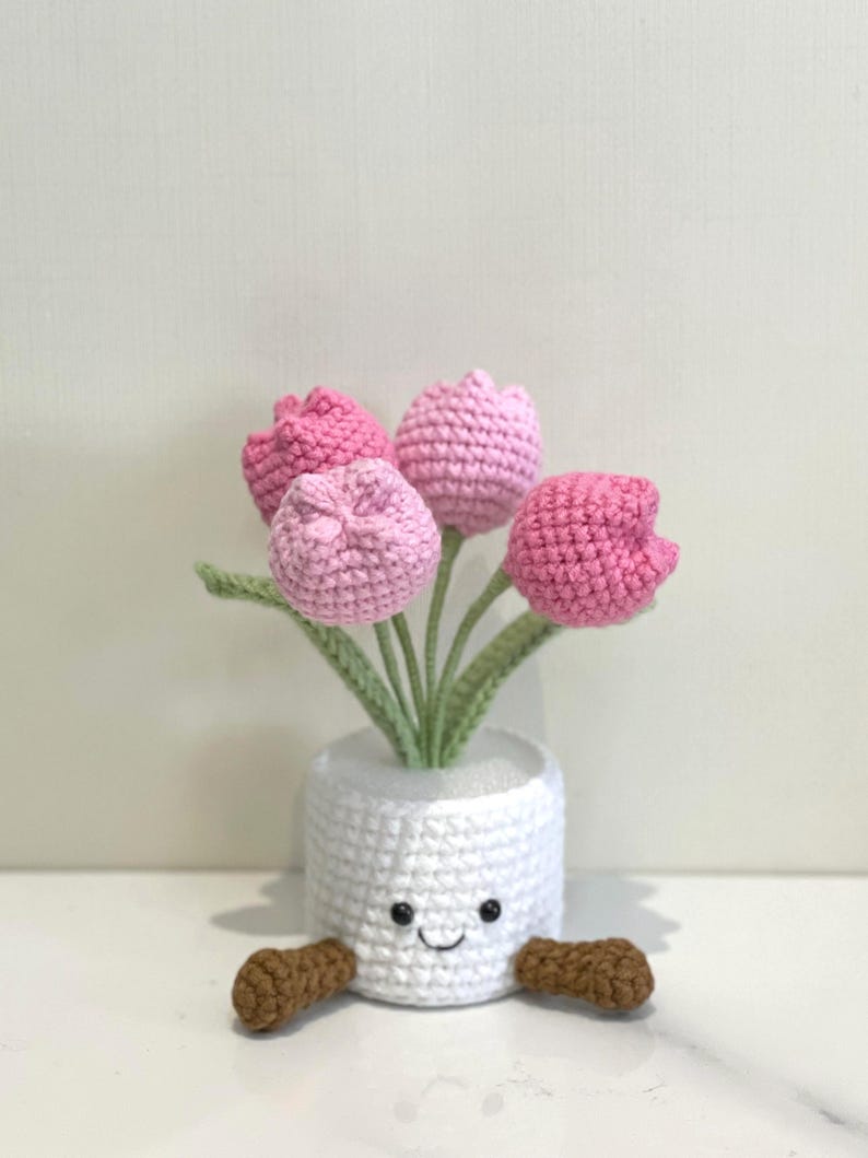 May include: A crocheted flower pot with a smiling face and brown feet, holding pink and light pink crocheted tulips with green stems and leaves. The white pot has a textured appearance.