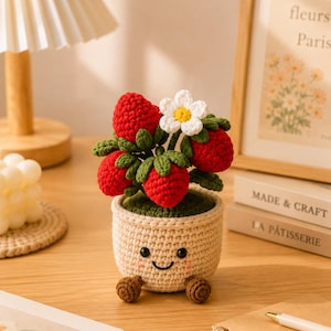 Peut inclure: Une plante fraise au crochet dans un pot beige avec un visage souriant. La plante présente des fraises rouge vif, des feuilles vertes et une fleur blanche avec un cœur jaune. Le pot possède deux petits pieds marron et une texture en laine.