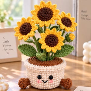 Könnte beinhalten: Eine gehäkelte Sonnenblumenpflanze in einem beigen Topf mit lächelndem Gesicht, rosa Wangen und zwei kleinen braunen Füßen. Die Pflanze besteht aus drei großen gelben Sonnenblumen mit dunkelbraunen Mitten, grünen Blättern und einer Knospe.