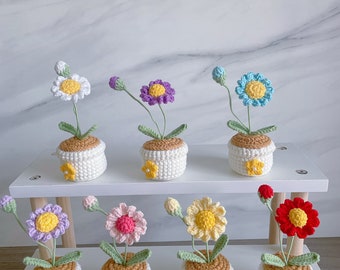 Puede incluir: Una colección de flores de ganchillo hechas a mano en pequeñas macetas blancas. Cada flor es de un color diferente, incluyendo blanco, morado, azul, rosa, amarillo y rojo. Las macetas tienen una parte superior marrón y un acento de flor amarilla. Las flores se exhiben en un estante blanco.