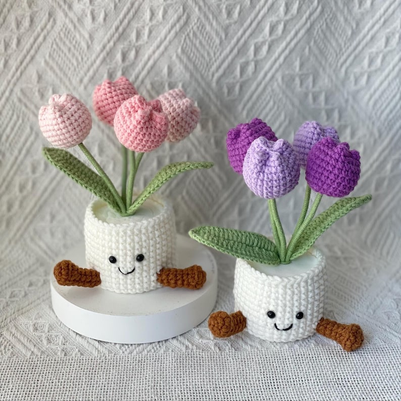 May include: Two crochet tulip flower arrangements in white pots with smiling faces and brown feet. One arrangement features pink tulips, the other purple. Green leaves and stems complete the handmade floral decor.
