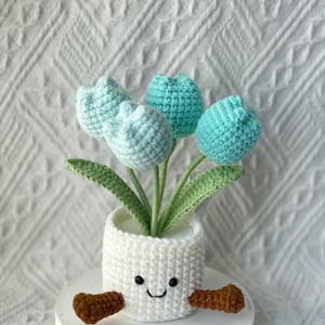 May include: A crocheted flower pot with a smiling face, holding four blue and light blue crocheted tulips. The pot is white, with brown arms. The flowers have green stems and leaves.