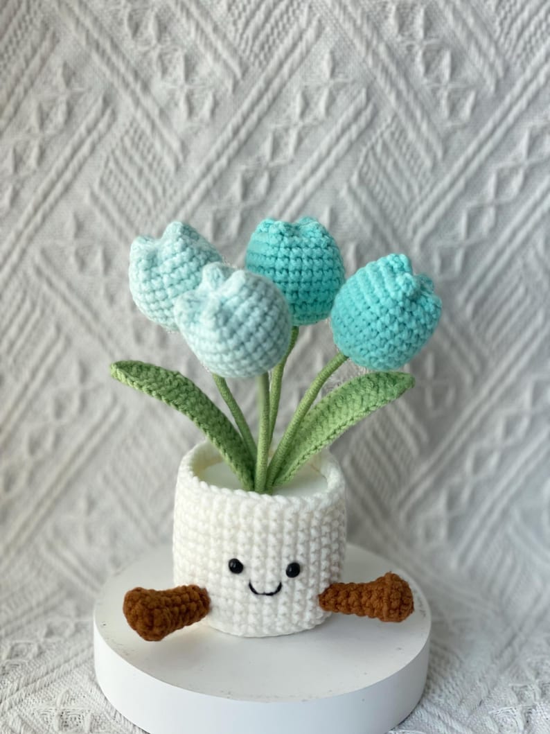 May include: A crocheted flower pot with a smiling face, holding four blue and light blue crocheted tulips. The pot is white, with brown arms. The flowers have green stems and leaves.